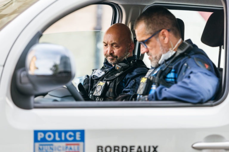 Deux policiers en uniforme sont assis à l'intérieur d'un véhicule de police blanc. Les deux policiers regardent vers le bas, lisant ou vérifiant peut-être quelque chose sur un appareil. Une étiquette « Police Municipale de Bordeaux » est visible sur la portière, et le mot « Bordeaux » est écrit en dessous. L'arrière-plan est flou.