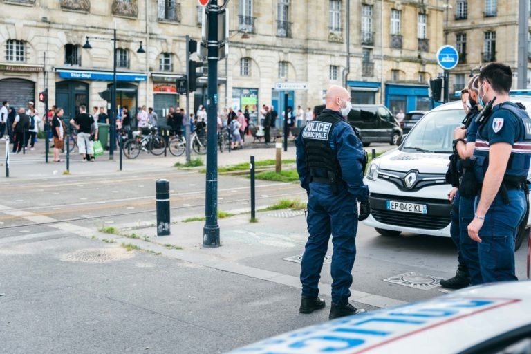 Trois policiers en uniforme et masqués de la police municipale de Bordeaux se tiennent à côté d'un véhicule de police blanc dans une rue. Un groupe de personnes est rassemblé à l'arrière-plan près de commerces et d'une ligne de tramway. Les bâtiments à l'arrière-plan sont en pierre claire, mettant en valeur un style architectural européen.