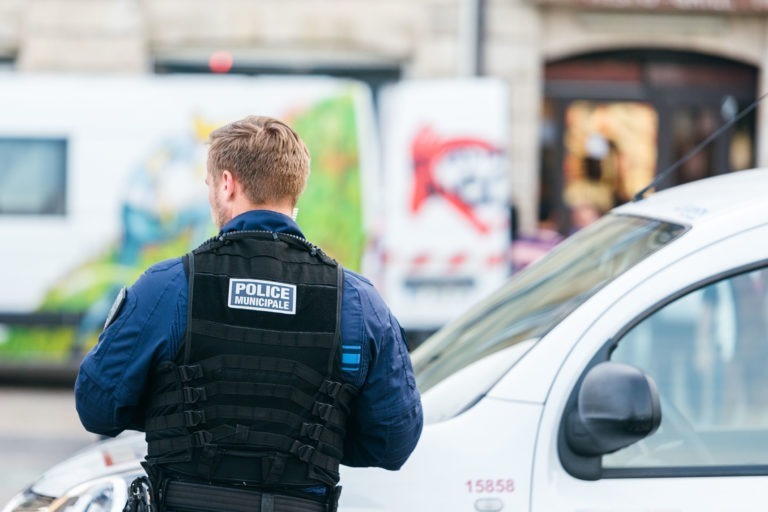 Un policier portant un gilet tactique noir marqué « Police Municipale » se tient à côté d'un véhicule de police blanc. L'agent, membre de la Police Municipale de Bordeaux, tourne le dos à la caméra. En arrière-plan, des peintures murales colorées ornent des fourgons blancs et une architecture légèrement floue complète la scène.