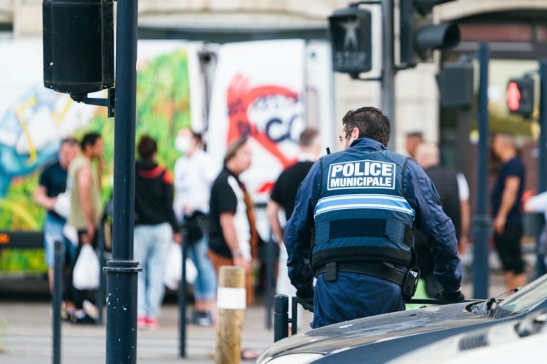 Un policier municipal portant un uniforme bleu marine avec une étiquette « POLICE MUNICIPALE » dans le dos et l'insigne distinctif de la police municipale de Bordeaux se tient à côté d'une voiture de patrouille dans une zone urbaine. En arrière-plan, plusieurs personnes sont rassemblées près d'un food truck avec des graffitis vibrants. Des feux de circulation et des poteaux de rue sont également visibles.