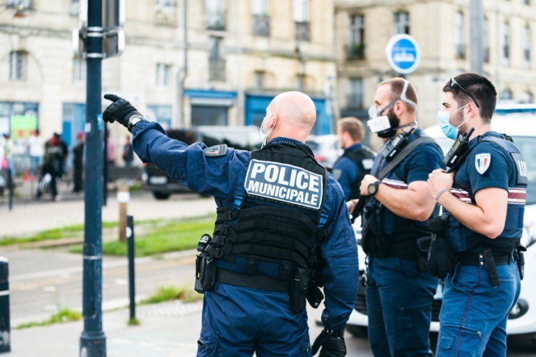 Trois agents de la police municipale de Bordeaux, en uniforme bleu et gilet de protection, se tiennent dans la rue. L'un des agents pointe vers l'avant, tandis que les deux autres regardent dans la même direction. Tous portent des masques. En arrière-plan, on voit des bâtiments, des piétons et un véhicule de police stationné.