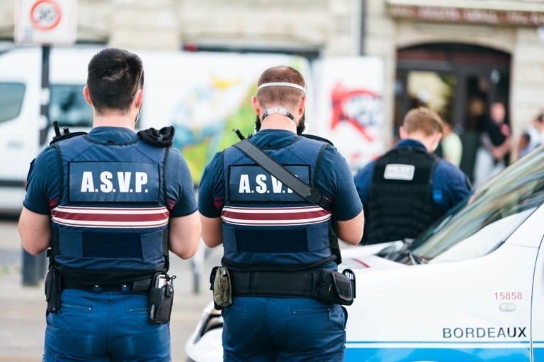 Trois agents de l'ASVP portant des uniformes bleu foncé et des gilets de protection sont vus de dos alors qu'ils se tiennent près d'un véhicule de la police municipale de Bordeaux. Ils se trouvent dans une zone urbaine avec des bâtiments flous et une camionnette en arrière-plan. Les agents sont concentrés sur une tâche, peut-être l'écriture ou la vérification d'appareils.