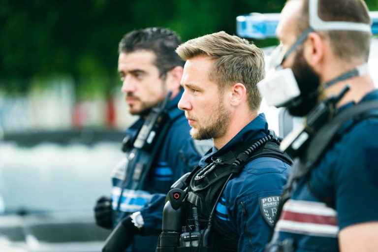 Trois agents de la police municipale de Bordeaux sont alignés. Chacun porte un uniforme sombre à rayures bleues et divers équipements, dont des caméras corporelles et des appareils de communication. L'homme au milieu est bien net, regardant fixement devant lui. L'arrière-plan est flou, avec de la verdure et des formes indistinctes.