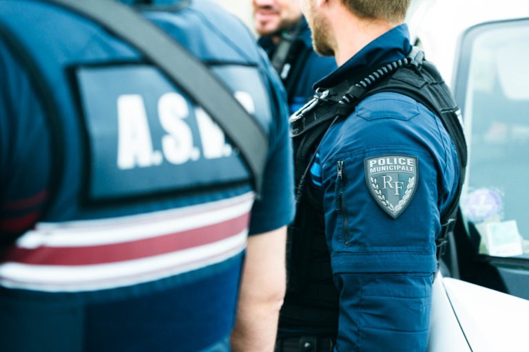 Trois policiers municipaux en uniforme bleu discutent près d'un véhicule. Au premier plan, on voit le dos d'un policier avec l'inscription « A.S.N.B » sur son uniforme. L'écusson de manche de l'agent central indique « Police Municipale RF ». Un autre agent partiellement visible et souriant discute avec le groupe, représentant la police municipale de Bordeaux.