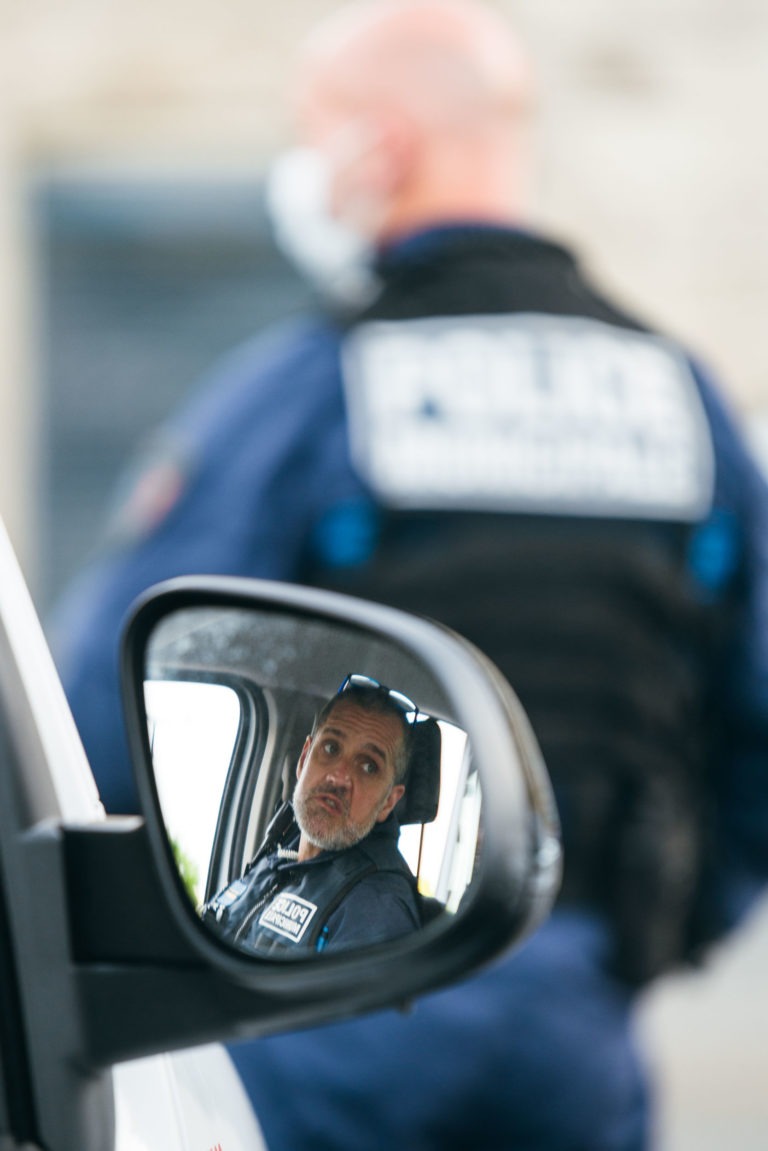 Un policier en uniforme se reflète dans le rétroviseur latéral d'un véhicule et regarde la caméra. Derrière le véhicule, un autre policier de la police municipale de Bordeaux, flou, se tient debout, le dos tourné, avec "Police Municipale" visible sur son gilet. Les deux policiers semblent être à l'extérieur en plein jour.