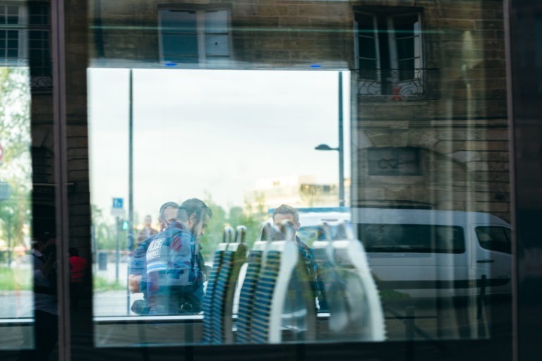 Le reflet dans une vitre montre trois personnes en mouvement et des meubles à l'intérieur, avec l'arrière-plan capturant une scène urbaine extérieure avec des bâtiments et une camionnette blanche garée. L'image a une qualité stratifiée, légèrement abstraite en raison des reflets et de la transparence du verre, presque comme si elle était vue à travers les yeux de la police municipale de Bordeaux.