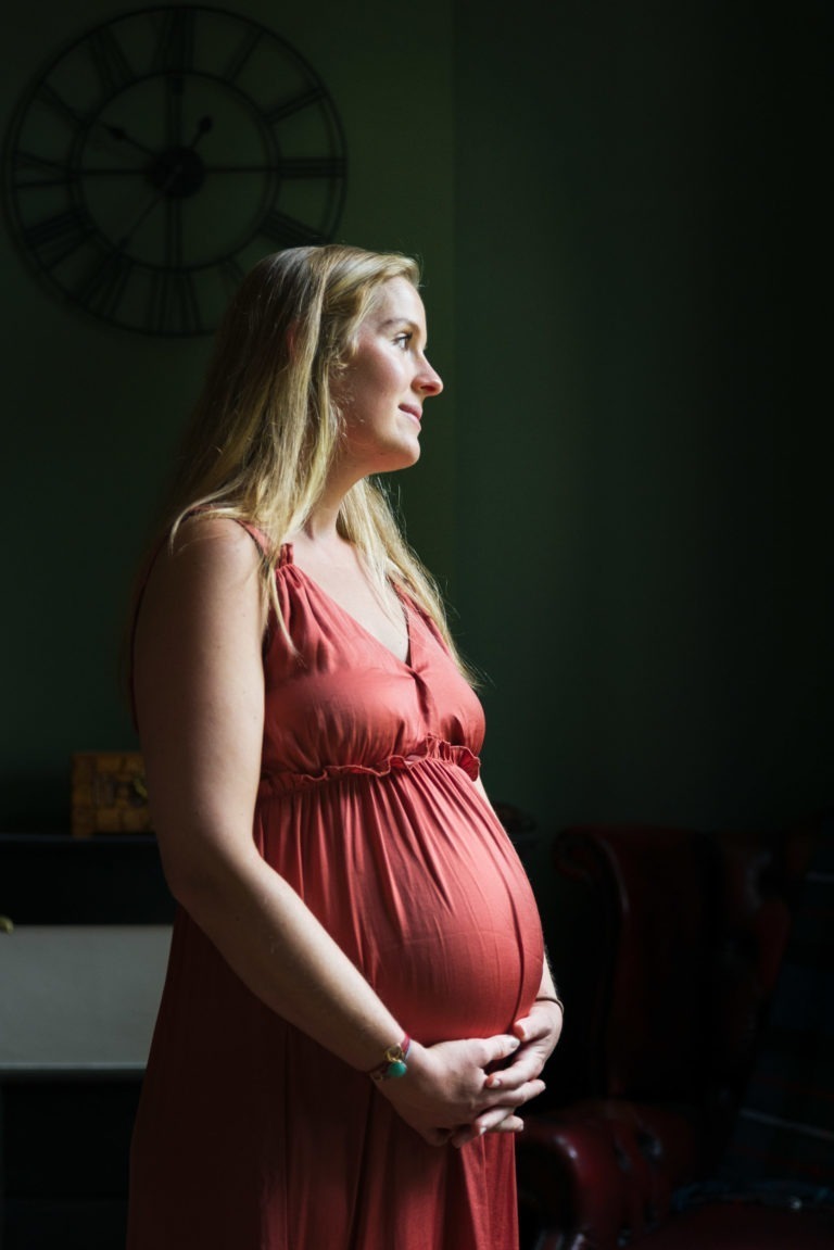 Une femme enceinte se tient de profil, le regard tourné vers la droite, dans une pièce faiblement éclairée au mur vert foncé. Elle porte une robe sans manches de couleur corail et pose ses mains sur son ventre. Une grande horloge murale est visible en arrière-plan, marquant l'attente naissance bébé. L'éclairage met en valeur son expression paisible et ses cheveux blonds.