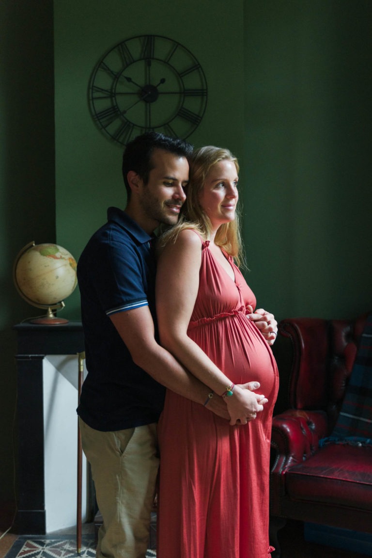 Une femme enceinte en robe corail se tient devant un fauteuil en cuir rouge. Un homme en chemise bleu marine et pantalon beige se tient derrière elle, tenant doucement son ventre. Ils ont tous deux l'air satisfaits en attendant la naissance du bébé. Un globe et une horloge sont visibles en arrière-plan, ajoutant une touche vintage à la pièce.