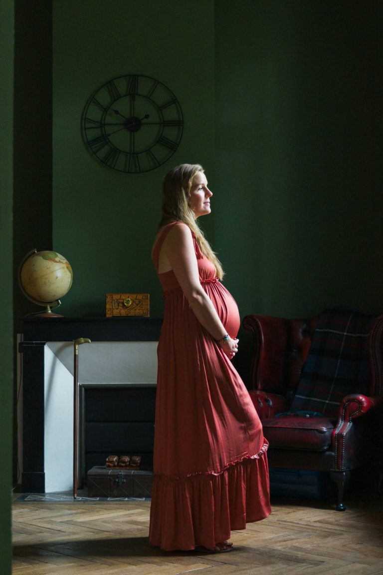 Une femme enceinte, vêtue d'une longue robe corail, se tient dans une pièce aux murs vert foncé. Elle pose ses mains sur son ventre rond et regarde vers la droite, loin de la caméra, perdue dans ses pensées sur l'attente de la naissance. La pièce est ornée d'une horloge vintage accrochée au mur, d'un globe terrestre sur le manteau de la cheminée et d'un fauteuil marron avec une couverture à carreaux.