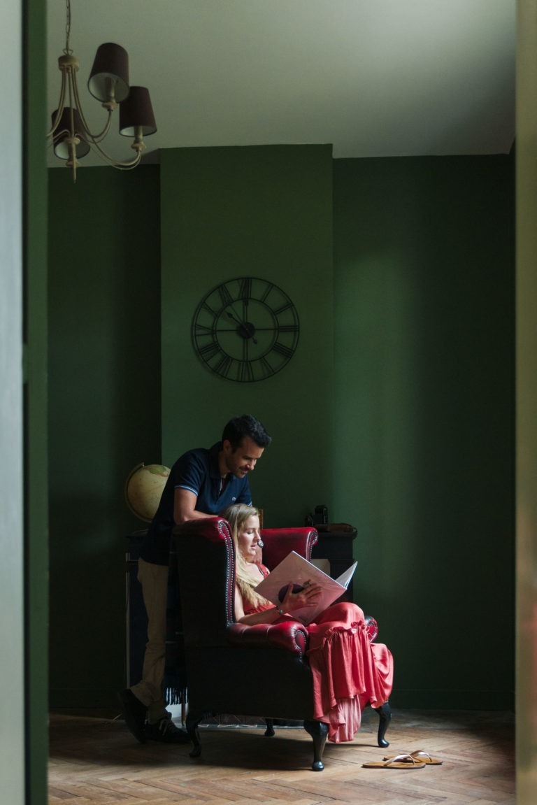 Un homme se tient debout derrière une jeune fille assise dans un fauteuil rouge moelleux, lisant ensemble un livre. La pièce a des murs verts, un globe terrestre sur un meuble et une grande horloge murale en arrière-plan. Le tout est éclairé par une douce lumière naturelle provenant d'une fenêtre voisine, créant une atmosphère remplie d'attente sereine.