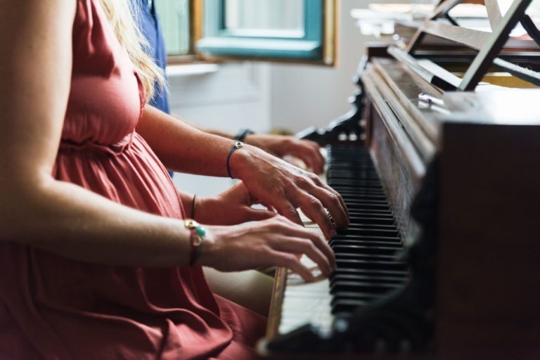 Gros plan sur deux personnes jouant du piano ensemble, attendant l'arrivée de leur bébé. La personne à gauche porte une robe rose et ses mains sont visibles sur les touches. La personne à droite, en chemise bleue à manches longues, a également les mains sur les touches. La lumière filtre à travers une fenêtre ouverte à proximité.