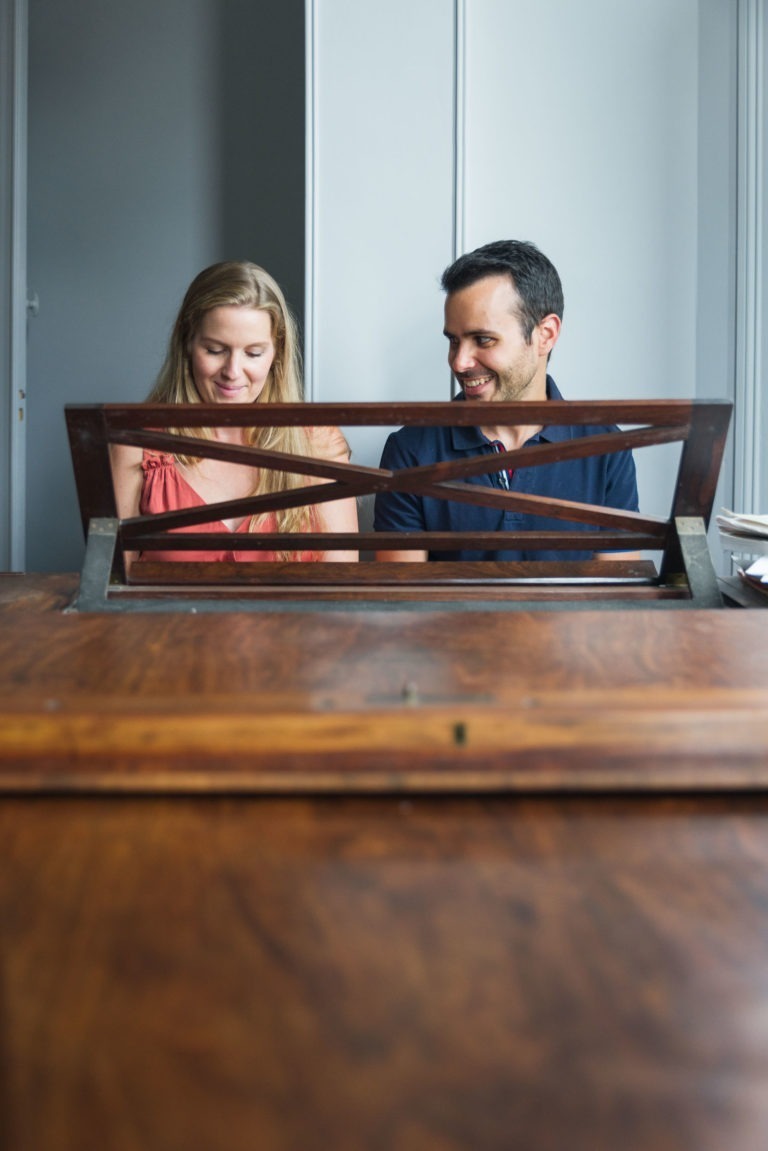 Une femme et un homme sont assis côte à côte devant un piano en bois dont le couvercle est partiellement ouvert, révélant le pupitre du piano. La femme, qui attend la naissance de son bébé, a de longs cheveux bruns et porte un haut rose sans manches, tandis que l'homme a les cheveux noirs courts et porte une chemise de couleur sombre. Tous deux sourient.