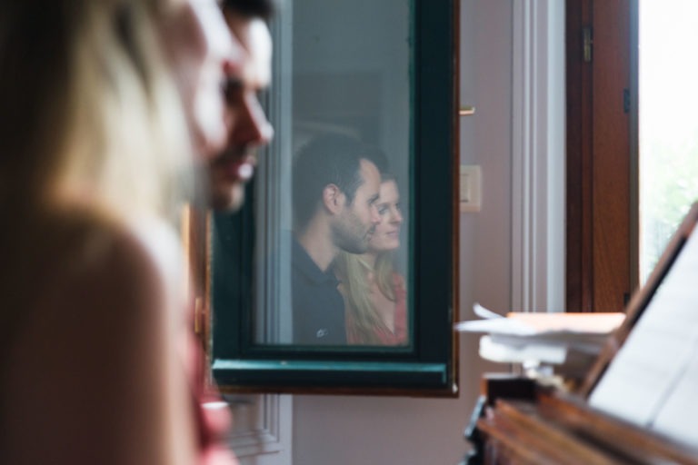 Gros plan d'un homme et d'une femme reflétés dans une petite fenêtre, partiellement masqués par une silhouette floue au premier plan. Ils regardent tous deux vers la droite avec une expression d'attente, et l'arrière-plan comprend un cadre de fenêtre en bois et une petite pile de papiers sur le côté droit de l'image.