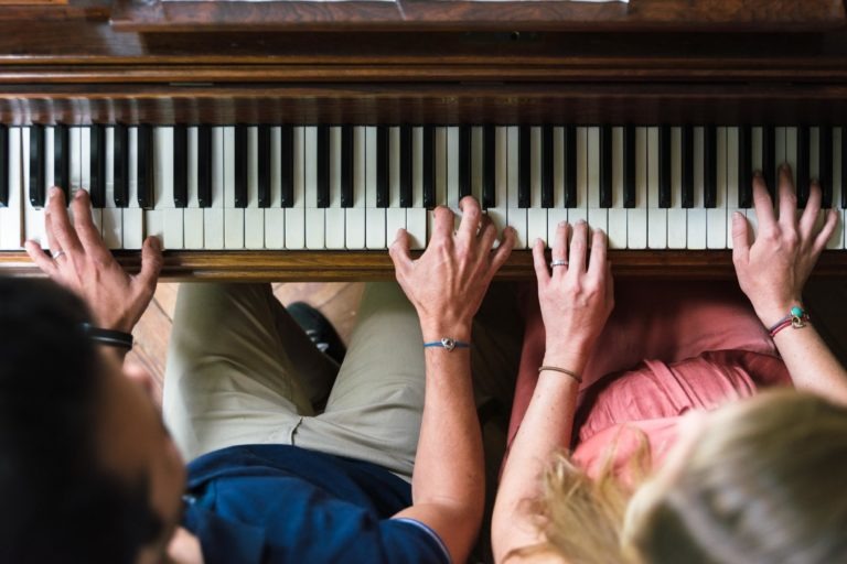 Vue de dessus de deux personnes jouant ensemble sur un piano en bois. La personne de gauche, qui attend peut-être la naissance de son bébé, porte une chemise bleue et un pantalon beige, tandis que la personne de droite porte une robe rose. Leurs mains sont positionnées sur les touches pour un duo, des bracelets ornant leurs poignets.