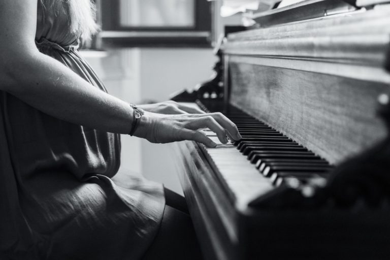 Photographie en noir et blanc montrant une personne assise à un piano droit, jouant un accord. Seuls son torse et ses mains sont visibles. La personne porte une robe et un bracelet au poignet gauche. L'arrière-plan comprend une fenêtre floue, ajoutant à l'atmosphère intime de la scène d'attente naissance bébé.