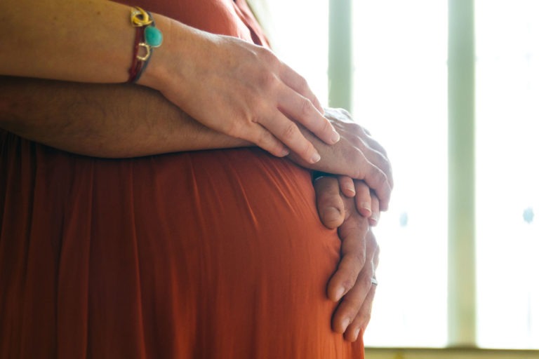 Gros plan d'une femme enceinte en robe orange, les deux mains posées délicatement sur son ventre, symbolisant l'attention et l'attente de la naissance du bébé. Sa main gauche et celle d'un partenaire sont visibles, elle portant un bracelet. L'arrière-plan est doucement éclairé par une fenêtre, créant une atmosphère sereine et chaleureuse.