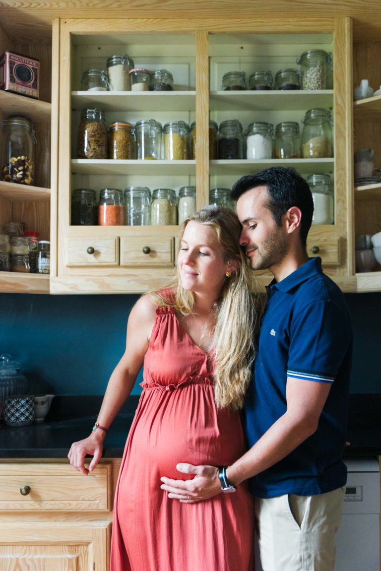 Une femme enceinte en robe corail se tient debout dans une cuisine, appuyée contre un meuble en bois rempli de bocaux et de récipients. À côté d'elle, un homme en polo bleu marine, la main sur le ventre, la regarde affectueusement tandis qu'ils partagent la joie d'attendre la naissance de bébé. Tous deux ont les cheveux clairs et la cuisine est dotée de murs bleus et de plans de travail en bois.