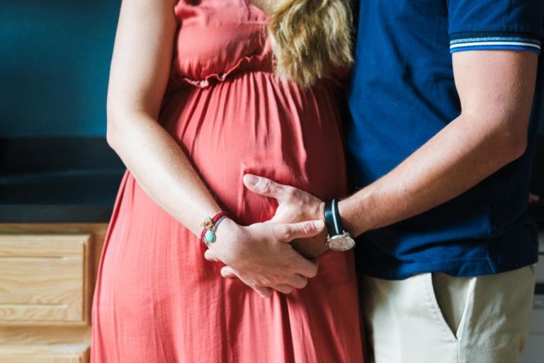 Gros plan d'une femme enceinte dans une robe corail sans manches, debout à côté d'un homme en chemise bleu marine et pantalon beige, tous deux berçant son ventre rond avec impatience pour la naissance de bébé. Ses longs cheveux blonds ondulés tombent sur un côté. Une armoire en bois est visible à l'arrière-plan.