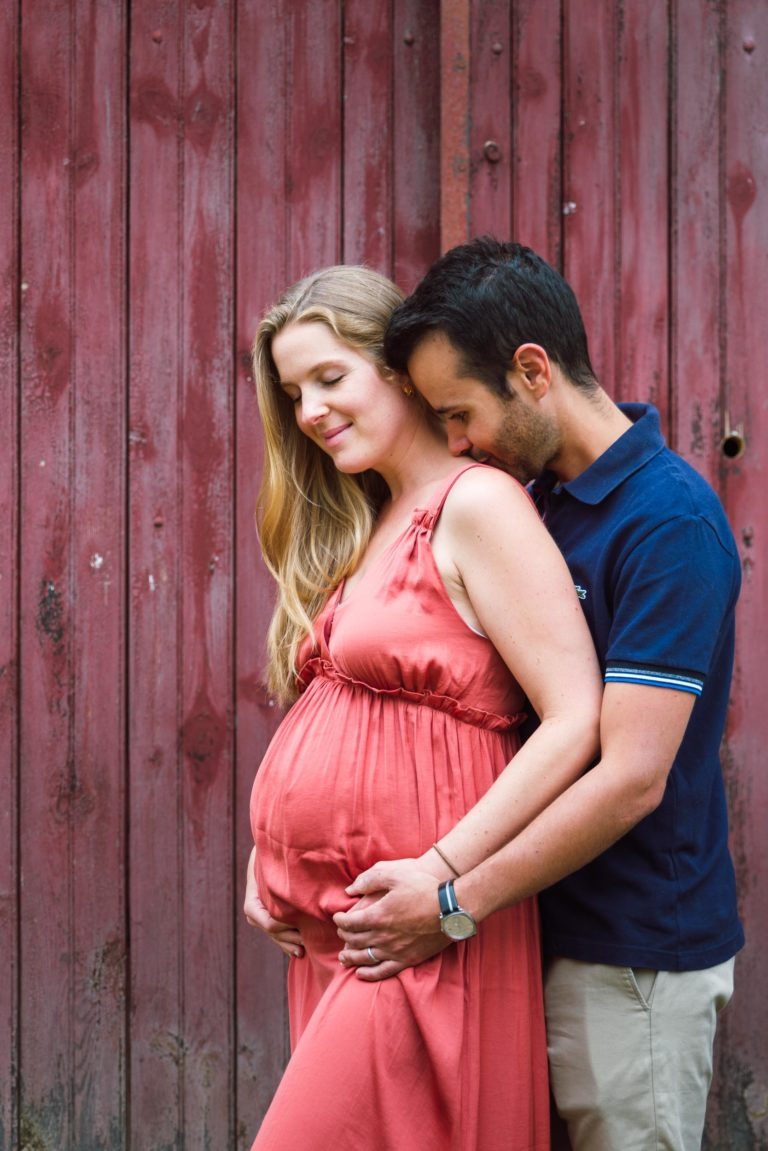 Une femme enceinte en robe corail se tient devant un mur en bois rouge rustique avec son partenaire, qui porte un polo bleu marine et un pantalon beige. Il se tient juste derrière elle, berçant son ventre rond avec ses deux mains tout en posant sa joue sur sa tête. Tous deux semblent satisfaits et paisibles, attendant la naissance de bébé.