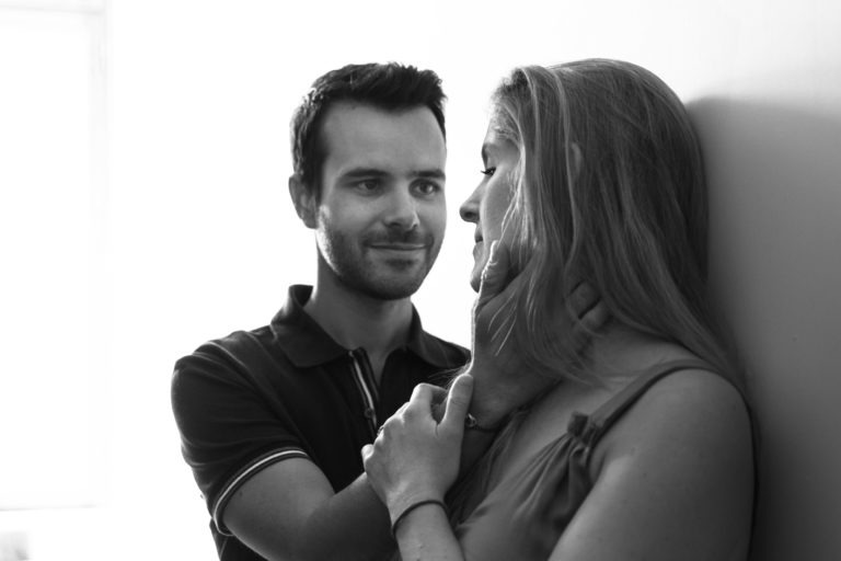 Photographie en noir et blanc d'un homme et d'une femme. L'homme, souriant doucement, a les cheveux courts et foncés et porte un polo foncé. Il tient doucement le visage de la femme avec ses deux mains. La femme, aux cheveux longs et clairs et portant un haut sans manches, le regarde avec une expression sereine alors qu'ils se tiennent debout sur un fond clair en attendant la naissance de leur bébé.