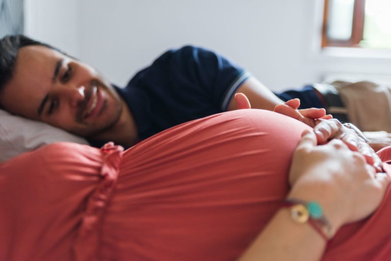 Un homme est allongé sur un lit, souriant et reposant sa tête sur un oreiller, face à une femme enceinte. Sa main repose doucement sur son ventre rond, recouvert d'une robe rose. Les mains de la femme sont également posées sur son ventre, portant un bracelet avec des perles bleues et dorées. Une fenêtre est visible en arrière-plan, capturant leur sereine attente naissance bébé.