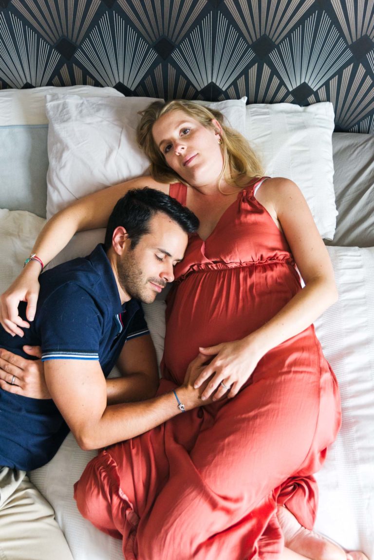 Une femme enceinte en robe orange est allongée sur un lit à la tête de lit à motifs géométriques, tenant son ventre en attendant la naissance de leur bébé. Un homme en chemise bleu marine est allongé à côté d'elle, la tête posée sur son ventre, la serrant dans ses bras. Tous deux semblent détendus et satisfaits. Des coussins blancs les entourent.