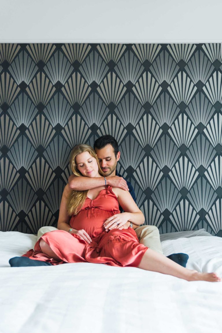 Une femme enceinte vêtue d'une robe rouge fluide et un homme en chemise noire sont assis sur un lit. L'homme enlace la femme par derrière tandis qu'elle se penche en arrière vers lui. Ils sont devant un mur à motifs en éventail, dégageant une aura de tranquillité détendue pendant leur attente de la naissance du bébé.