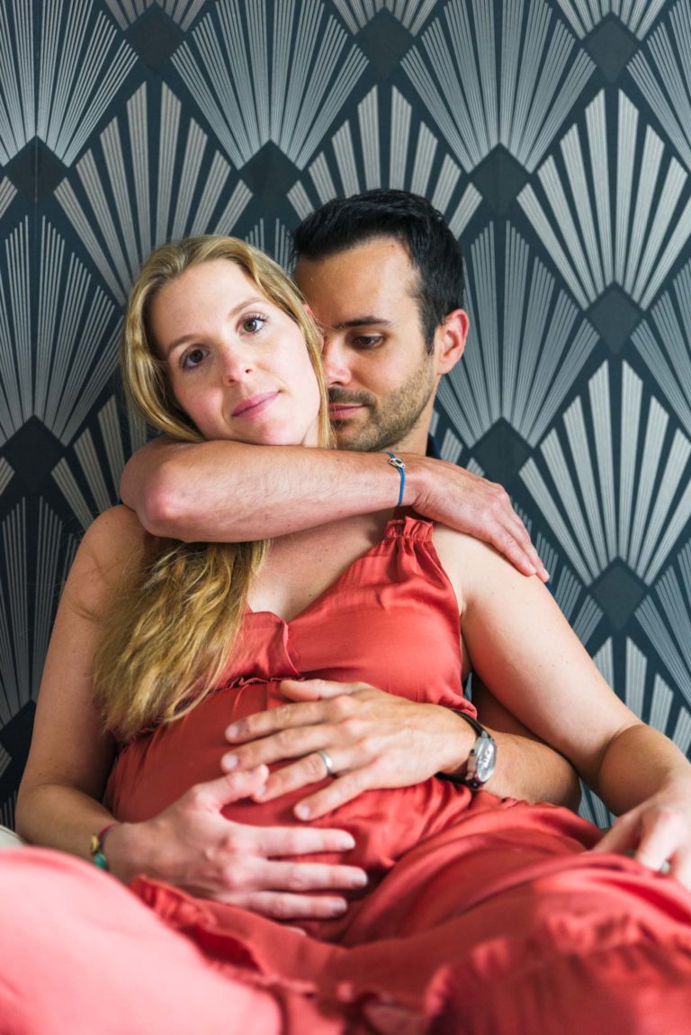 Un homme enlace une femme enceinte par derrière, assis contre un mur au motif art déco, incarnant la beauté sereine de l'attente de la naissance. La femme, vêtue d'une robe corail sans manches, a de longs cheveux blonds et pose ses mains sur son ventre. L'homme, vêtu d'un haut sans manches, l'entoure de ses bras avec un regard serein.