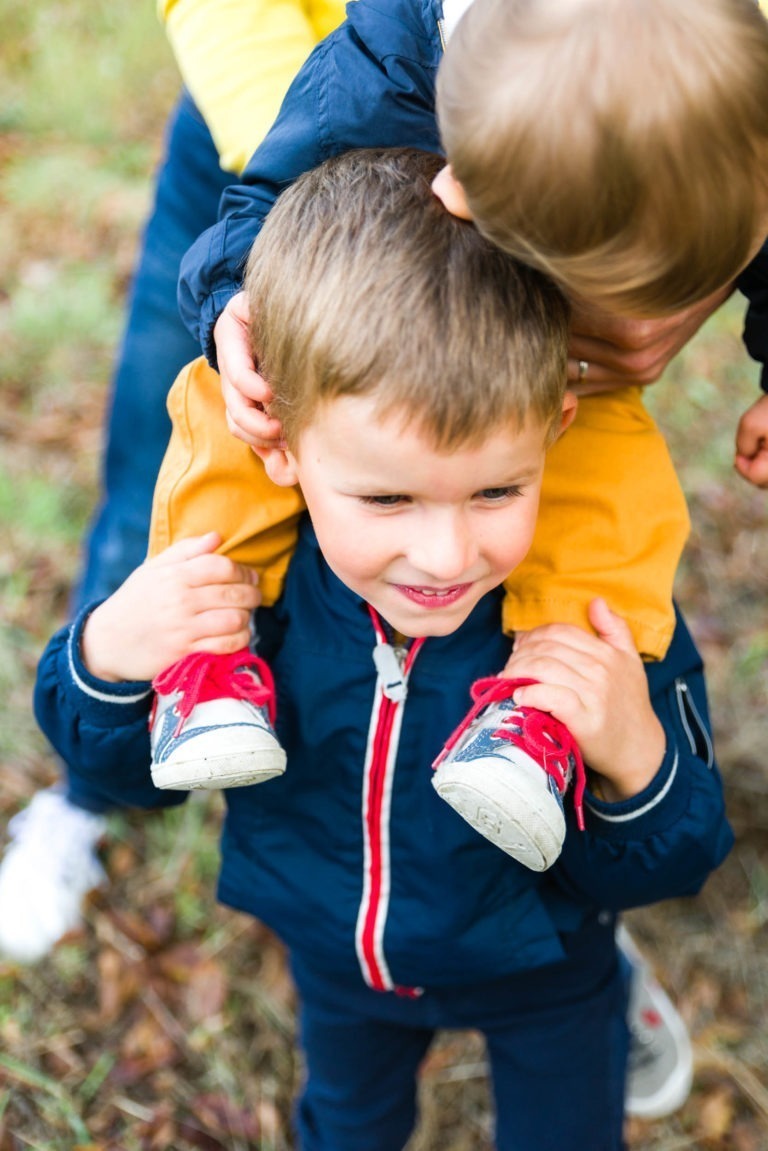 Un jeune garçon, vêtu d'une veste bleue à fermeture éclair rouge et d'un pantalon foncé, porte sur ses épaules un enfant plus jeune, lui aussi vêtu d'une veste bleue et d'un pantalon jaune moutarde. Le visage du plus jeune est partiellement visible, et tous deux sont dehors, sur le sol herbeux et feuillu, précieux souvenirs d'enfance de Bordeaux.