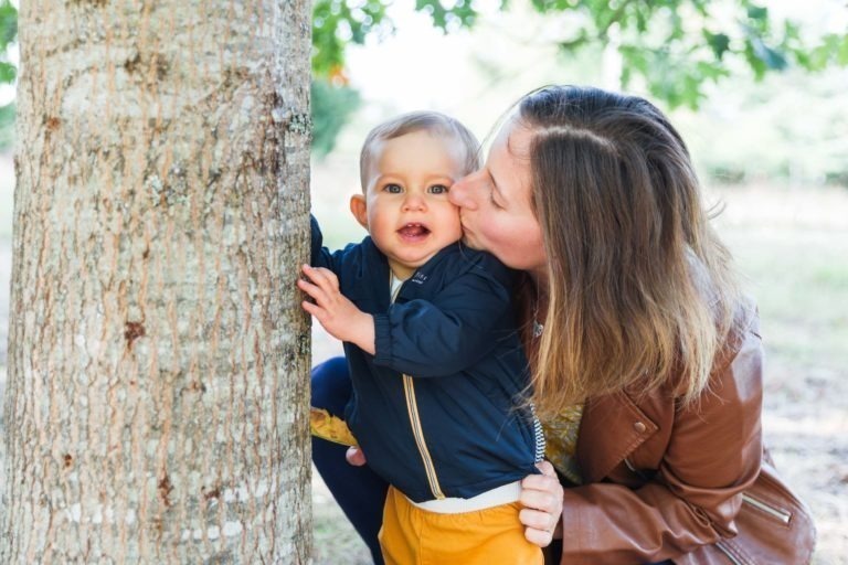 Une femme aux cheveux châtains, vêtue d'une veste marron, embrasse un bambin souriant aux cheveux blonds courts qui se tient debout et touche un arbre. L'enfant, vêtu d'une veste sombre et d'un pantalon jaune vif, incarne l'essence des souvenirs d'enfance au milieu du feuillage vert et de la douce lumière du cadre naturel extérieur de Bordeaux.