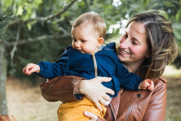 Une femme aux cheveux châtains mi-longs, vêtue d'une veste en cuir marron, tient dans ses bras un bébé souriant vêtu d'une veste bleu marine et d'un pantalon jaune moutarde. Ils sont en plein air à Bordeaux, entourés d'arbres et de verdure, profitant d'une agréable journée qui rappelle les souvenirs d'enfance.