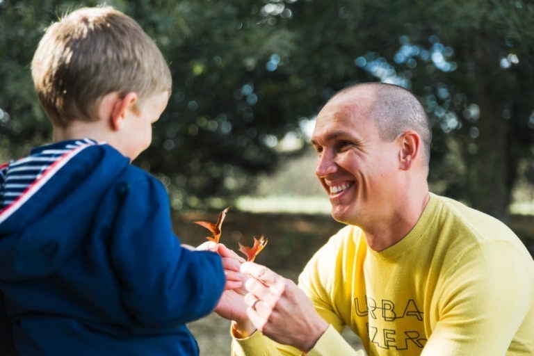 Un homme au crâne rasé sourit et tend des feuilles d'automne à un jeune garçon aux cheveux blonds courts, vêtu d'une veste bleue. Ils sont en extérieur à Bordeaux, entourés d'arbres verts et d'un soleil tacheté, créant une atmosphère chaleureuse et joyeuse. L'homme porte un pull jaune, évoquant des souvenirs d'enfance.