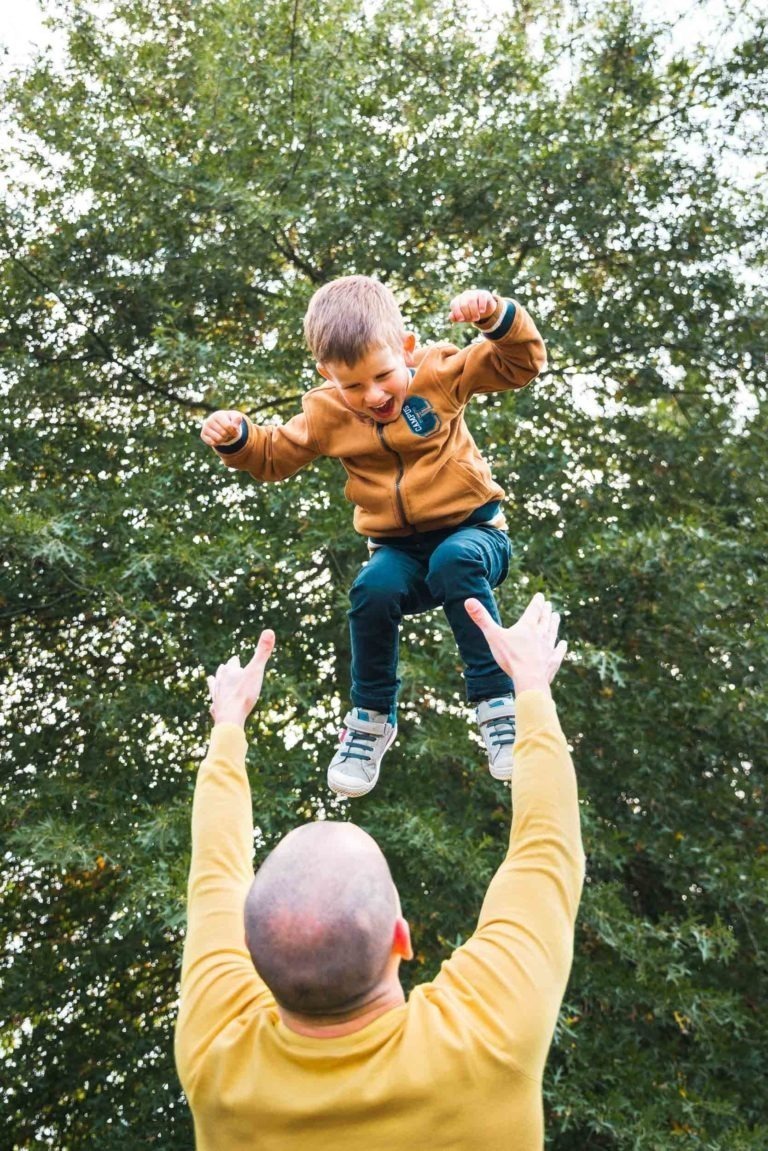 Un enfant joyeux, vêtu d'une veste marron et d'un pantalon vert, est lancé en l'air par un adulte au crâne rasé, qui porte un pull jaune. L'arrière-plan est orné d'arbres verts luxuriants, créant un décor extérieur vivant et dynamique rappelant les souvenirs d'enfance à Bordeaux.