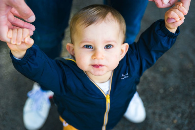 Un bambin aux cheveux châtain clair et aux yeux bleus regarde vers le haut tout en tenant les mains d'un adulte. L'enfant porte une veste bleu marine avec une fermeture éclair jaune et un pantalon bleu clair. Les jambes de l'adulte en jean bleu et chaussures blanches sont partiellement visibles en arrière-plan, rappelant les souvenirs d'enfance de Bordeaux, car la scène semble se dérouler en extérieur.