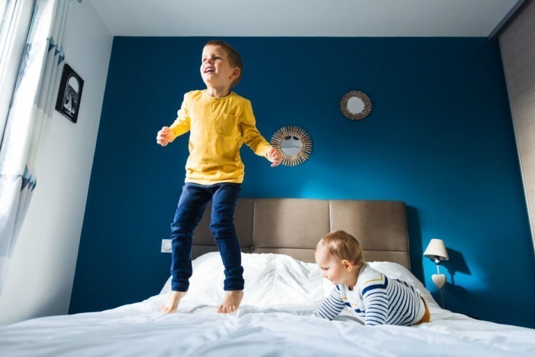 Deux jeunes enfants sont sur un lit dans une chambre au mur bleu rappelant les souvenirs d'enfance. L'enfant de gauche est debout et saute, vêtu d'une chemise jaune et d'un jean bleu. L'enfant de droite rampe et porte une chemise rayée. La pièce, décorée de deux miroirs ronds et d'une table de nuit avec une lampe, évoque des souvenirs de Bordeaux.