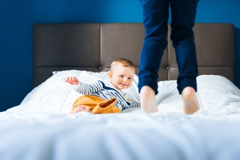 Un bébé portant une chemise rayée et un pantalon jaune est allongé sur un lit aux draps blancs. Un adulte en pantalon bleu foncé se tient au bord du lit, seules ses jambes étant visibles. L'arrière-plan présente un mur bleu et une tête de lit marron, évoquant les souvenirs d'enfance de Bordeaux, tandis que le bébé lève les yeux avec joie.