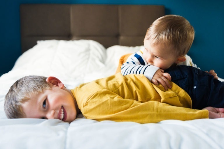 Deux jeunes enfants sur un lit, en train de se battre de manière ludique. L'aîné, vêtu d'une chemise jaune à manches longues, est allongé sur le ventre, souriant à la caméra. Le plus jeune, vêtu d'un pull rayé, est sur le dessus, enlaçant et mordant le dos de l'aîné de manière ludique. Cette scène de souvenirs d'enfance se déroule devant des draps blancs et une tête de lit sombre.