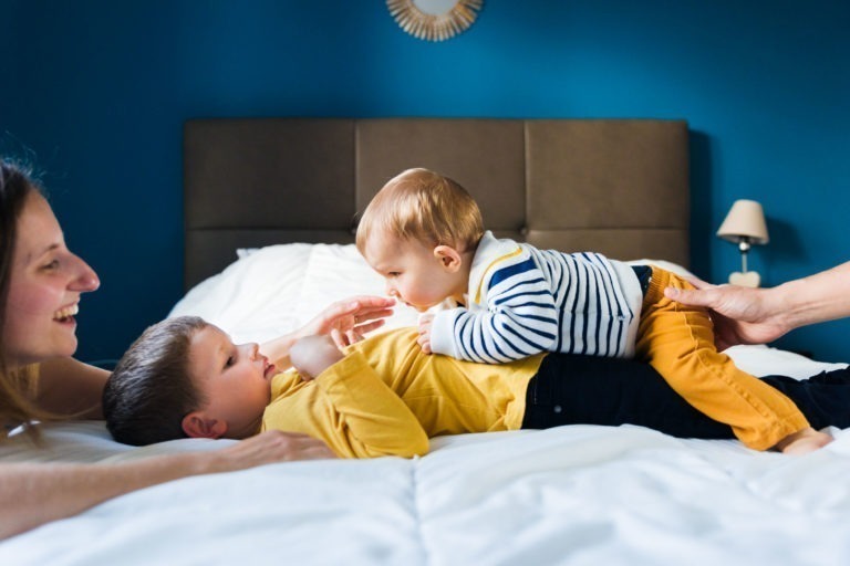 Une femme souriante est allongée sur un lit avec un jeune garçon rieur en chemise jaune et un bébé en pull rayé grimpant joyeusement sur lui. Une autre main soutient doucement le bébé. L'arrière-plan, rappelant les souvenirs d'enfance, présente une tête de lit marron et un mur bleu avec une petite lampe.