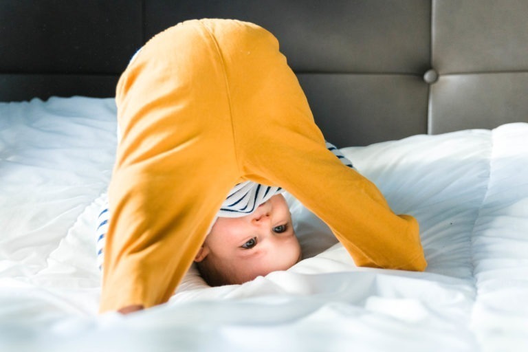 Un bébé portant un pantalon jaune et une chemise rayée joue sur un lit blanc, évoquant des souvenirs d'enfance. Le bébé est à l'envers, la tête et les mains posées sur le lit, regardant à travers ses jambes. L'arrière-plan comprend une tête de lit capitonnée sombre, rappelant un décor chaleureux de Bordeaux. La scène est éclairée par la lumière naturelle.