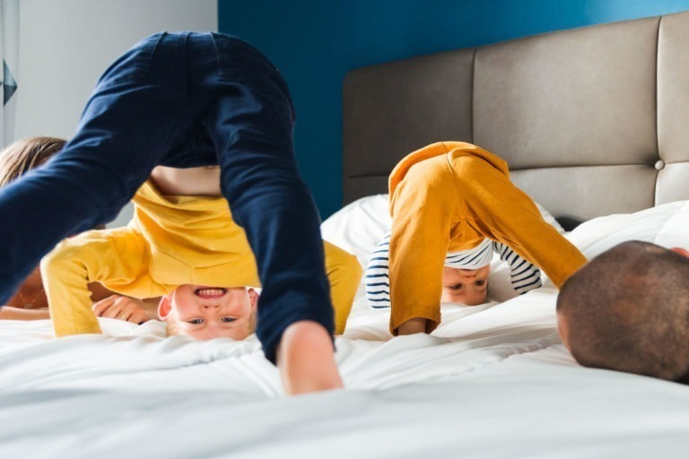 Deux jeunes enfants font le poirier sur un lit, souriant largement. L'un porte une chemise jaune avec un pantalon bleu, l'autre une chemise rayée avec un pantalon jaune. La tête d'un adulte est partiellement visible en bas à droite. Le lit a une tête de lit beige contre un mur bleu, évoquant des souvenirs d'enfance chéris.