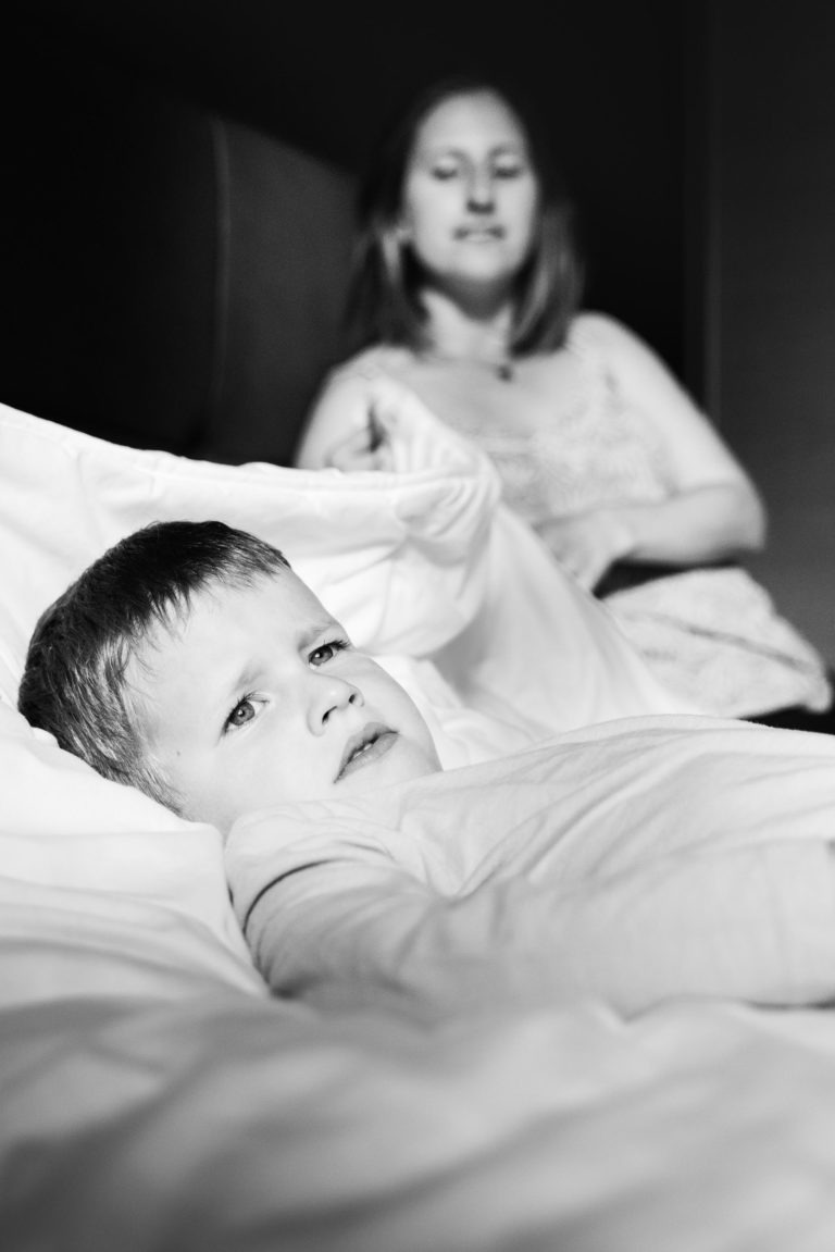 Une image en noir et blanc montre un jeune garçon allongé dans son lit, l'air inquiet. En arrière-plan, une femme aux cheveux mi-longs l'observe assise sur le lit. Des draps blancs recouvrent partiellement le garçon, et l'ambiance générale semble sérieuse et contemplative, évoquant les souvenirs d'enfance bordelaise.