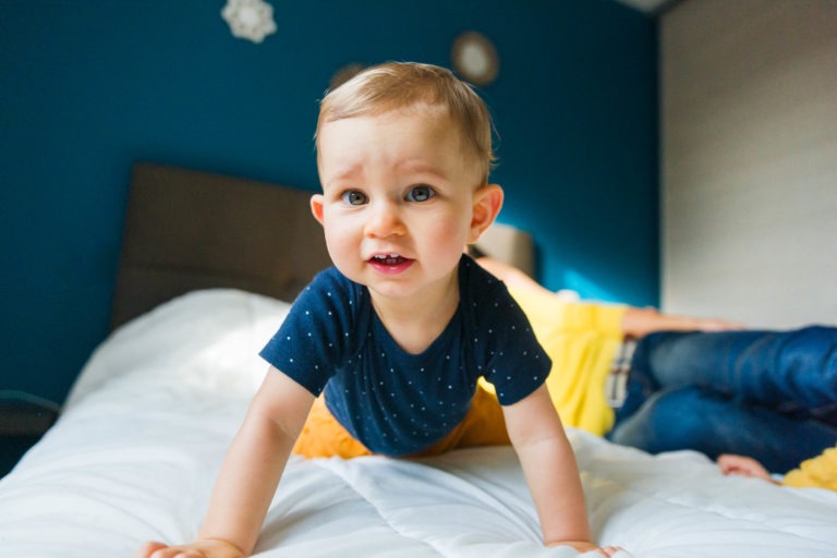 Un jeune enfant aux cheveux châtain clair, vêtu d'une chemise à manches courtes bleu foncé à petits pois blancs et d'un pantalon couleur moutarde, rampe sur une couette blanche. Le mur bleu sarcelle en arrière-plan présente un décor rond flou et une personne en chemise jaune en partie visible allongée, évoquant les souvenirs d'enfance de Bordeaux.