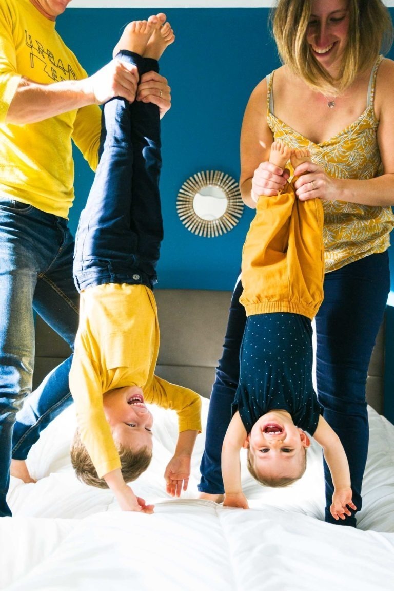 Deux adultes tiennent par les jambes deux bambins souriants sur un lit, la tête en bas, créant de joyeux souvenirs d'enfance. Les bambins, vêtus de hauts jaunes et noirs, rient joyeusement tandis que les adultes, souriants eux aussi en jeans bleus et hauts jaunes, profitent de ce moment ludique. Un mur bleu avec un miroir décoratif sert de toile de fond.