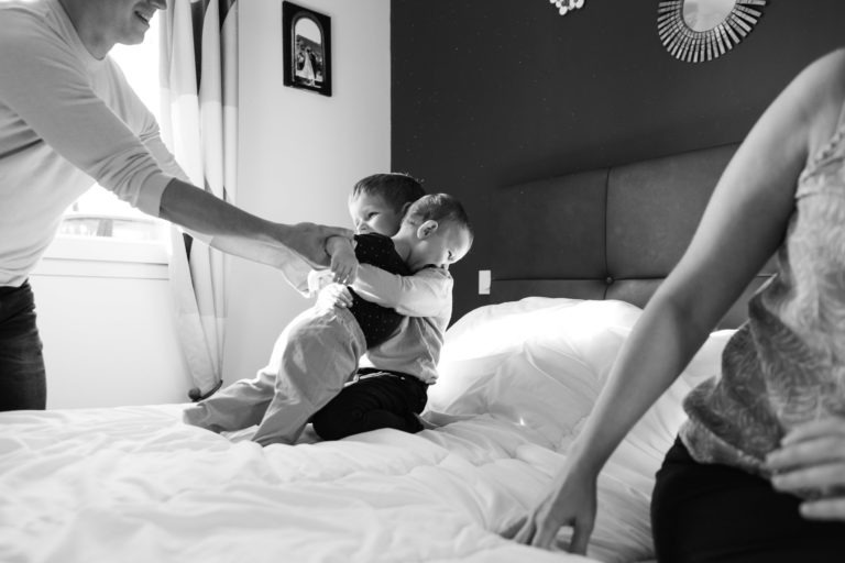 Une photo en noir et blanc montre un jeune enfant serrant dans ses bras un bébé sur un lit. Un adulte à gauche tient la main du bébé, tandis qu'un autre adulte à droite est légèrement hors cadre. La chambre de Bordeaux possède une grande tête de lit tapissée, une fenêtre avec des rideaux et quelques souvenirs d'enfance accrochés au mur.