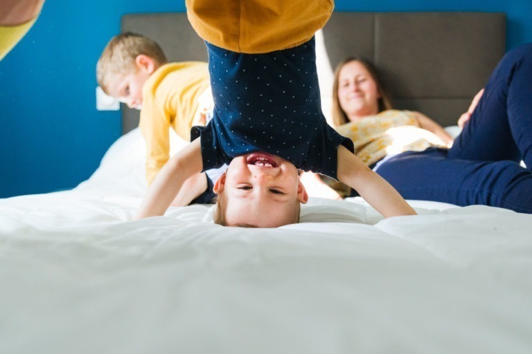 Un jeune enfant en chemise bleu marine et pantalon jaune est photographié la tête en bas, en train de faire le poirier sur un lit, tout sourire. En arrière-plan, un adulte et un autre enfant sont assis sur le lit, souriant et satisfaits. La pièce a des murs bleu vif et une atmosphère légère et joyeuse, évoquant les souvenirs d'enfance de Bordeaux.