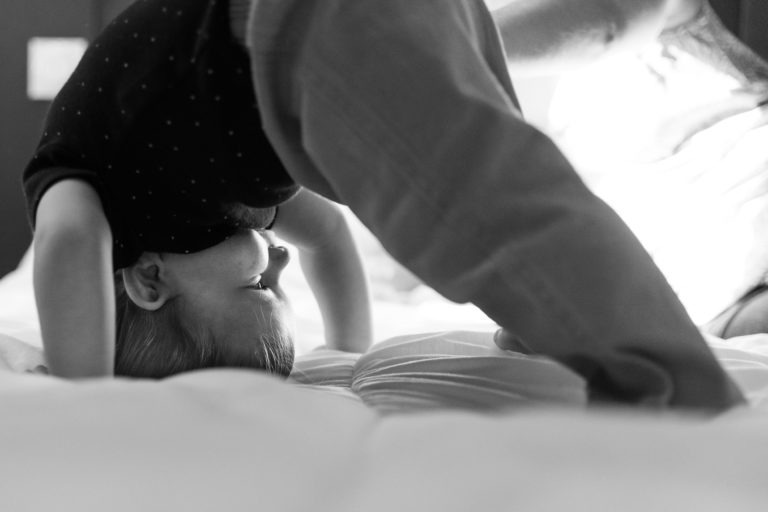 Photographie en noir et blanc d'un jeune enfant qui fait joyeusement le poirier sur un lit. L'enfant, vêtu d'une chemise et d'un pantalon sombres, est baigné d'une douce lumière venant du côté droit, soulignant son expression enjouée. L'arrière-plan flou met en valeur l'activité de l'enfant, évoquant des souvenirs d'enfance à Bordeaux.