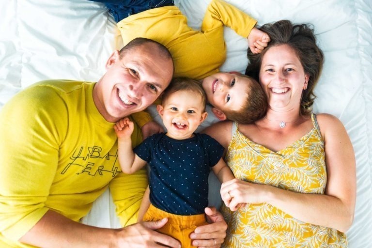 Une famille souriante de quatre personnes est allongée sur un lit. Le père, vêtu d'une chemise jaune, est à gauche, à côté d'un jeune bébé portant une chemise bleue. Un jeune enfant vêtu d'une chemise jaune est partiellement allongé sur la mère, qui porte une robe jaune à motifs blancs. Tous regardent l'objectif et sourient, capturant des souvenirs réconfortants d'enfance que Bordeaux chérirait.