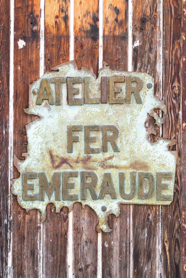 Une vieille enseigne en métal patiné aux bords rouillés est fixée sur un mur en bois. L'enseigne indique « ATELIER FER EMERAUDE » en lettres majuscules en relief, faisant allusion à la Ferronnerie d'Art Bordeaux. Le bois derrière l'enseigne est vieilli avec des planches verticales, montrant des signes d'usure et de vieillissement.