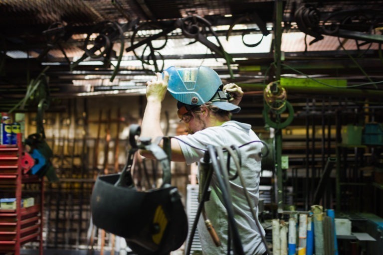 Un ouvrier en chemise blanche se trouve dans un atelier industriel, un casque bleu sur la tête. L'arrière-plan est encombré de divers outils, machines et équipements, notamment des câbles suspendus, une boîte à outils rouge et des tiges métalliques. La scène à la Ferronnerie d'Art Bordeaux est faiblement éclairée et animée.