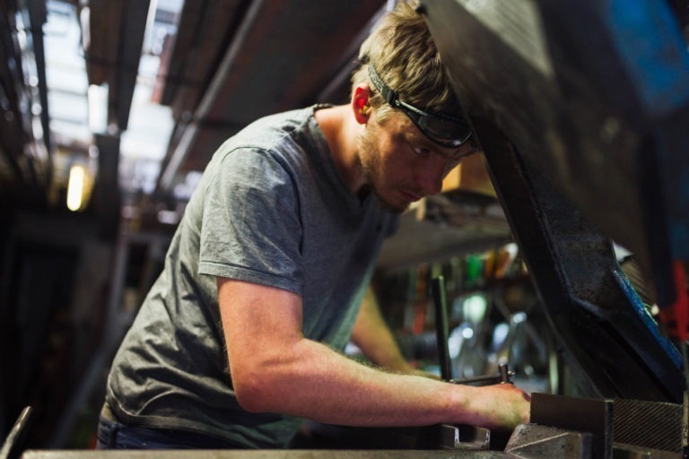 Un homme portant un t-shirt gris et des lunettes de sécurité travaille intensément sur des machines dans un environnement industriel avec divers composants métalliques. L'espace de travail est faiblement éclairé, ce qui met en valeur sa concentration alors qu'il se penche sur sa tâche. Des tuyaux et des équipements sont visibles en arrière-plan, rappelant l'artisanat de la Ferronnerie d'Art Bordeaux.